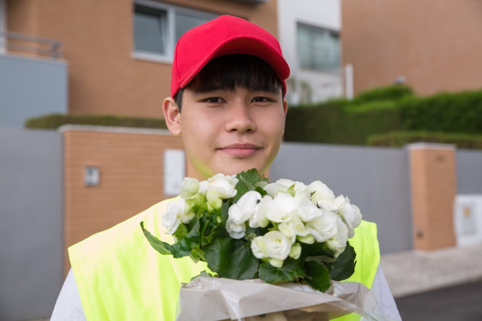 A young delivery person wearing a red cap and a high-visibility yellow vest is holding a bouquet of white flowers with green leaves, wrapped in white paper. The flowers are fresh, with soft petals and