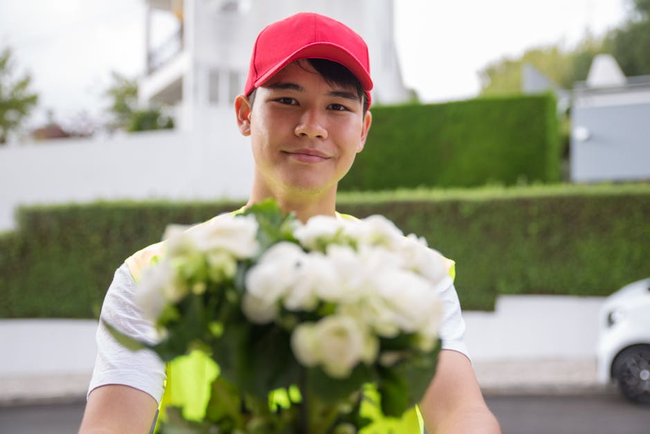 A delivery person wearing a bright pink cap and a high-visibility vest is smiling while holding a bouquet of fresh white flowers with green foliage. The bouquet features tightly arranged petals, givin