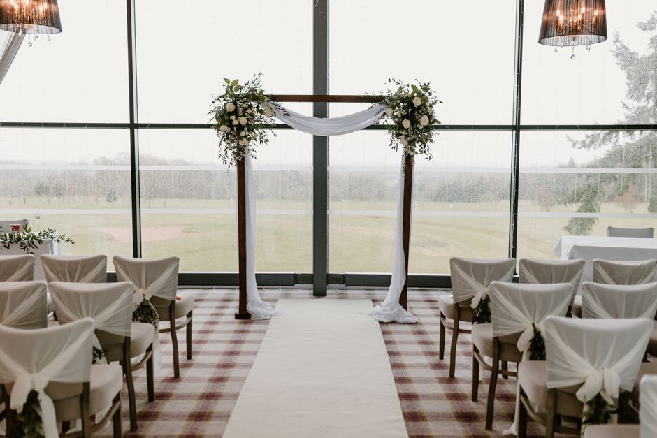 A flower arrangement featuring white roses and greenery decorates a wooden wedding arch, draped with white fabric, inside a modern venue with large floor-to-ceiling windows revealing an outdoor landsc