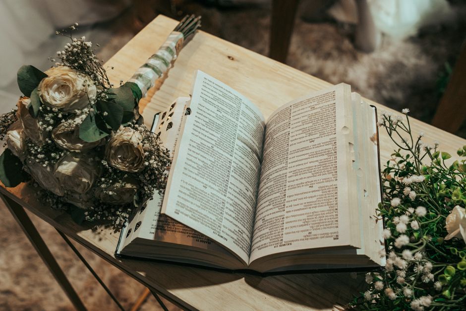 A bouquet of cream-colored roses and white baby's breath flowers, wrapped in green and white striped paper with a ribbon, is placed on a wooden table next to an open Bible. To the right, there is a se