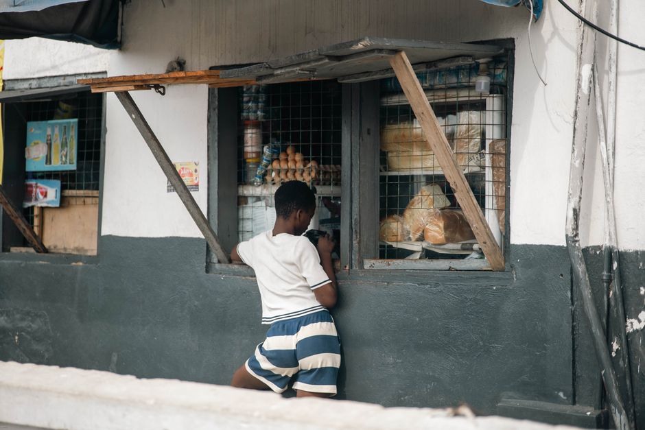 A young boy with dark skin, wearing a white t-shirt and blue-and-white striped shorts, stands in front of a small shop window that has wire mesh covering it. The window displays various baked goods an